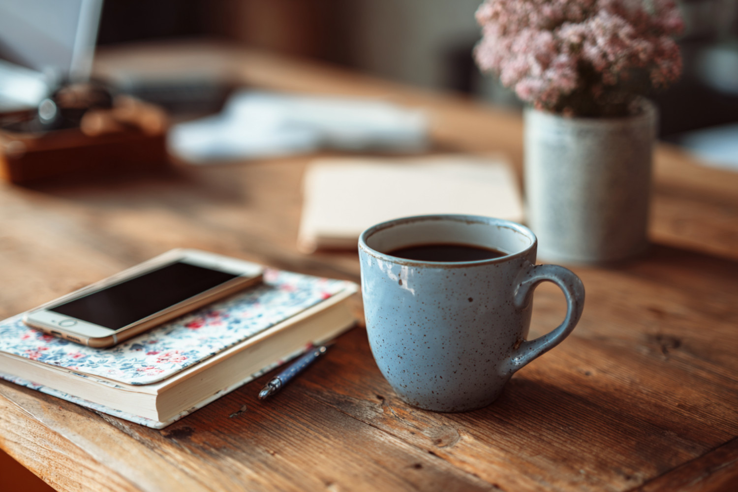 Eine blaue Tasse auf einem Tisch, daneben ein Notizbuch und darauf ein Smartphone